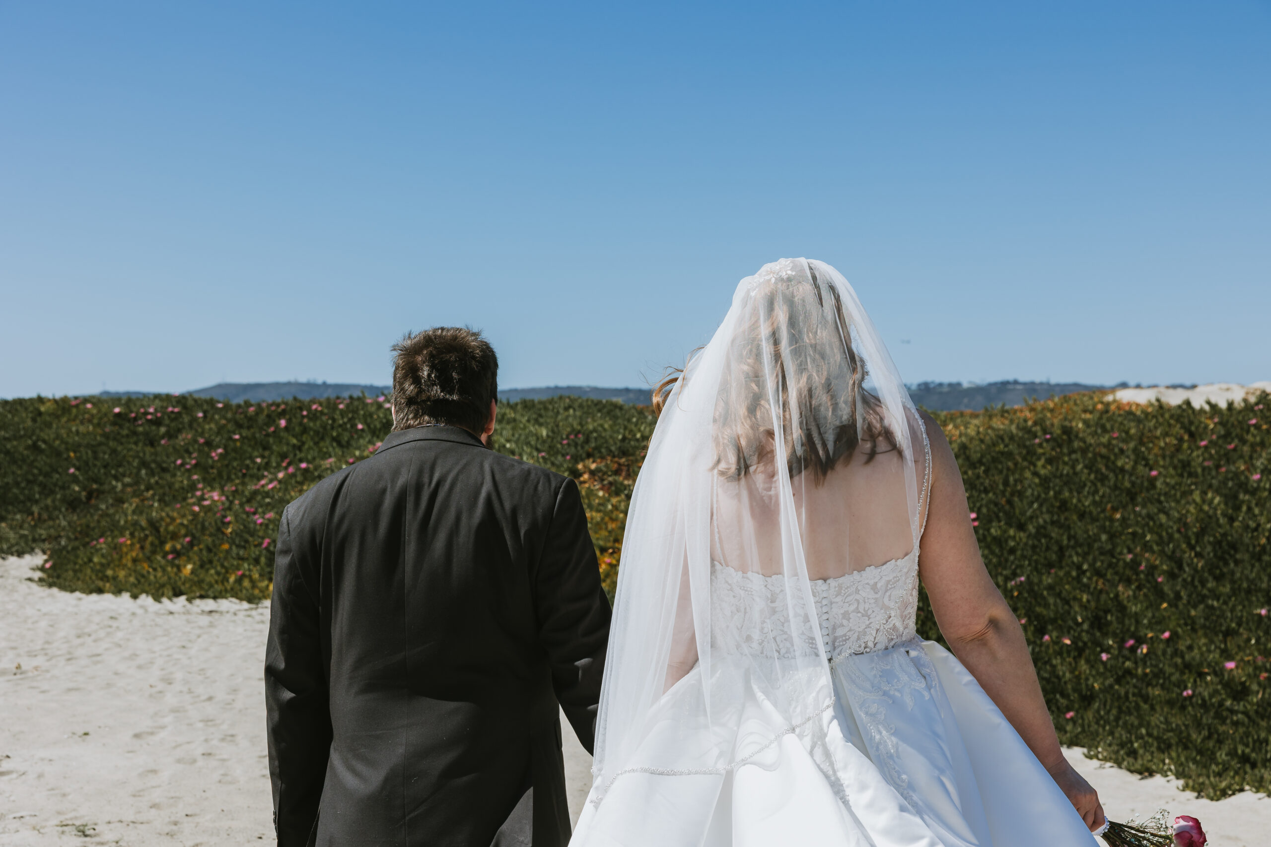 Photo is taken from behind a bride in a veil and a groom in a black suit walking on a beach with the sand dunes in the distance